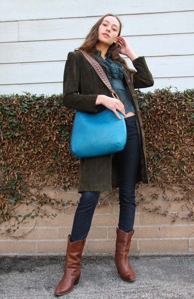 Beautiful woman standing in front of brick wall in jeans and olive green suede coat with teal lace ruffle blouse with peacock blue shoulder strap handbag