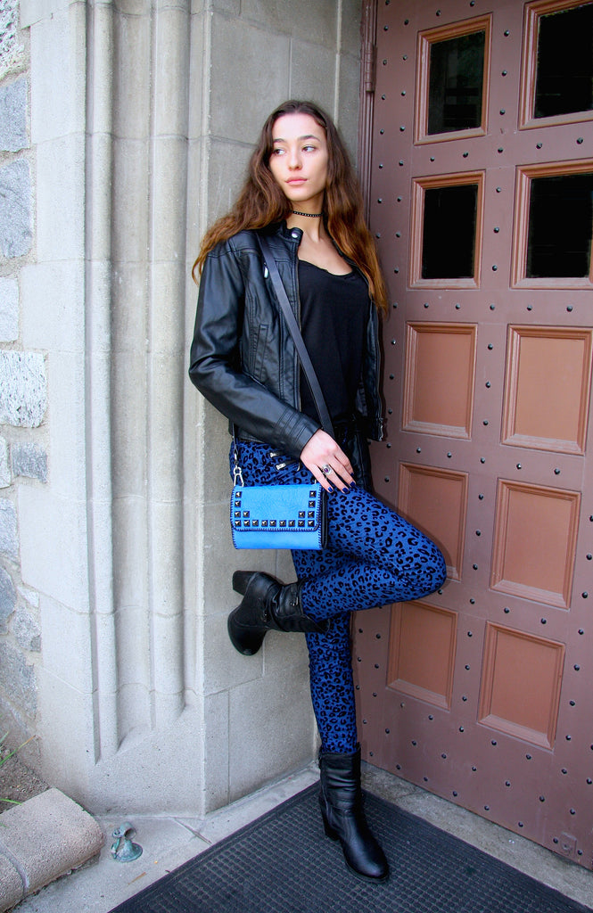 woman wearing blue leopard print pants and black leather jacket with blue leather studded cross body bag leaning in doorway 