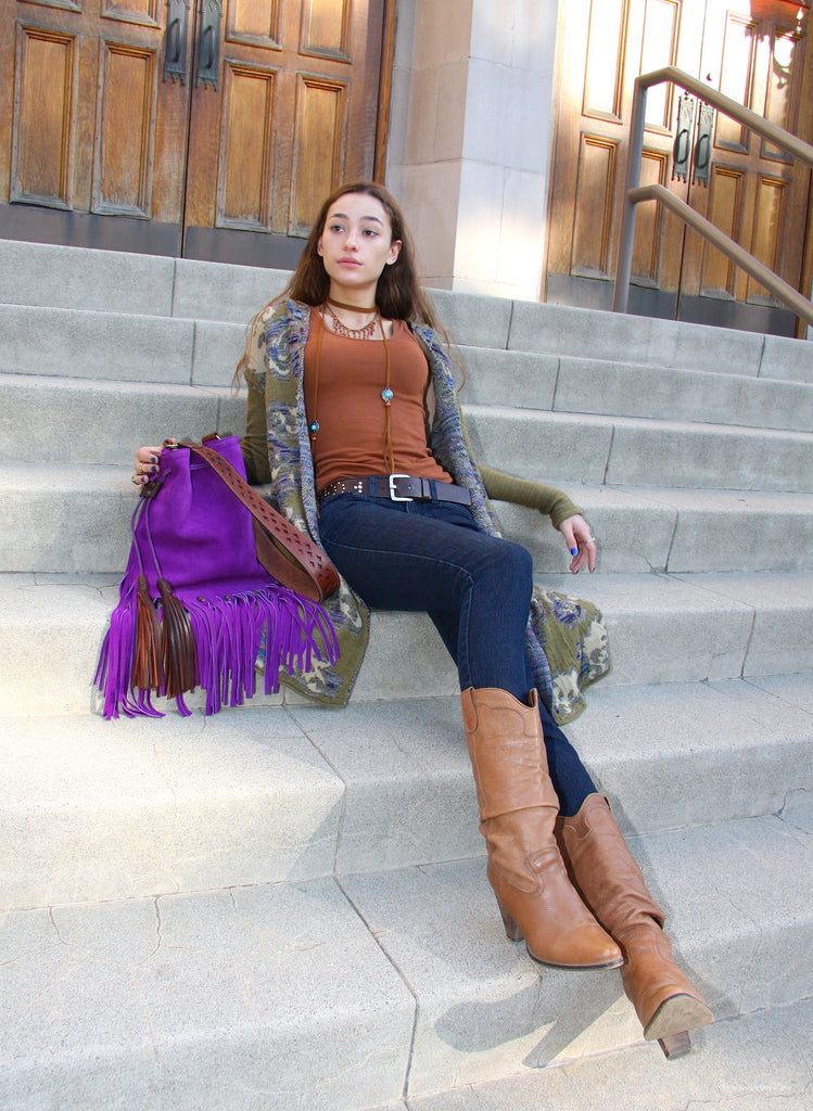 beautiful girl sitting on steps next to purple suede fringe drawstring handbag wearing jeans and a free people cardigan sweater