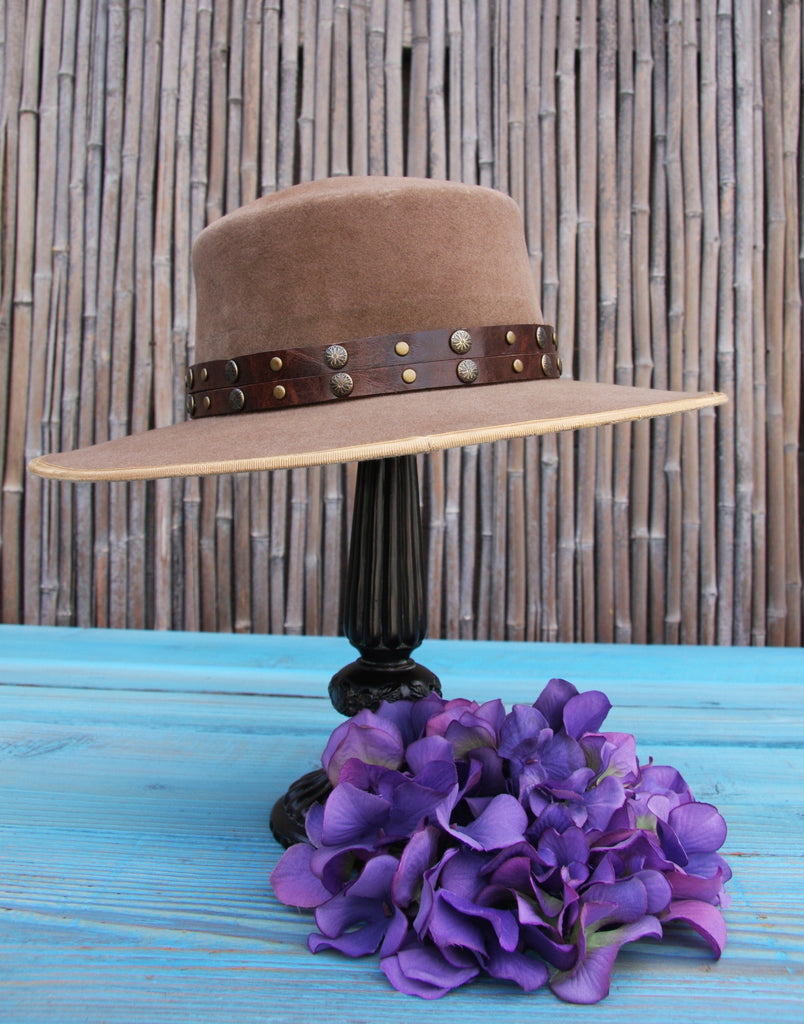 Tan bolero bohemian hat with brown chic hatband studded with antiqued brass studs sitting on a blue wood table with bamboo background
