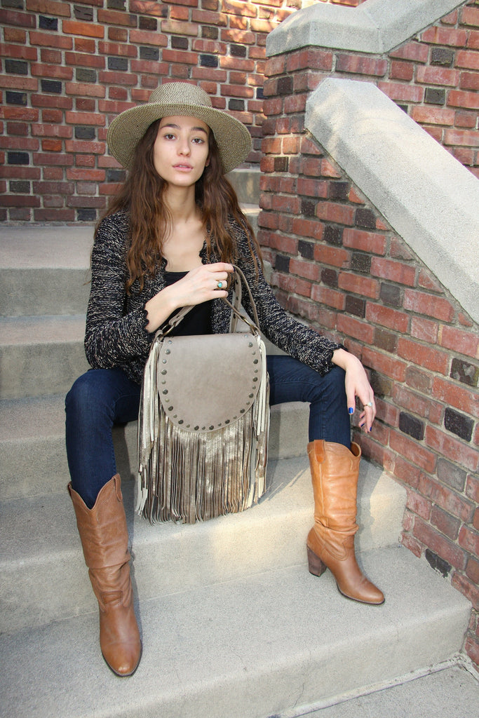 Beautiful woman sitting on stairs holding beige flap handbag with silver fringe and antiqued gold studs on flap wearing jeans cowboy boots and straw hat