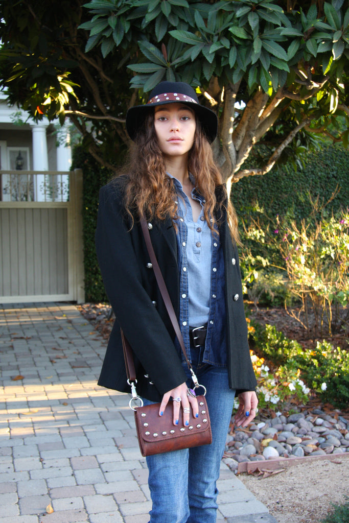 beautiful woman standing in garden wearing small brown studded crossbody handbag over black pea coat with jeans and black fedora 